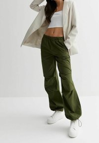 Zelené kalhoty s pružným pasem a volným střihem, vyrobené z lehké látky. Kombinovány s bílým crop topem a krémovým sakem, doplněné bílými teniskami.