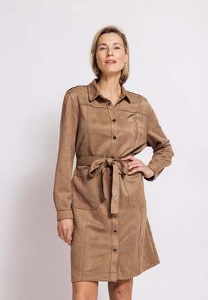 Woman wearing a brown buttoned shirt dress with a collar and tied belt, standing with one hand on her hip against a plain background.