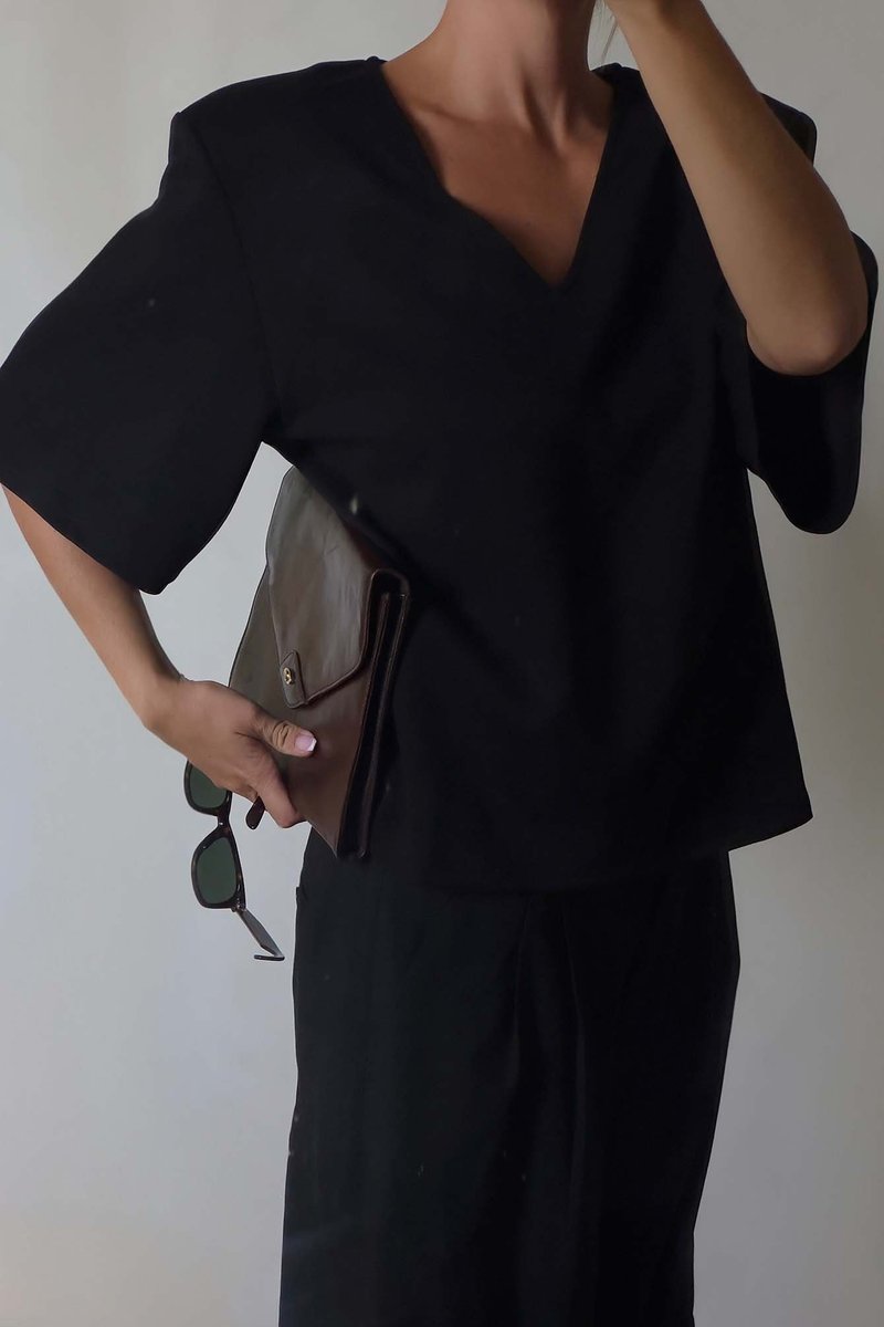 Mustade lühikeste varrukatega särk V-kaelusega, koos pruuni nahast portfelliga ja tumedate päikeseprillidega, neutraalse taustaga.
