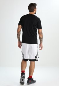 Mann mit Tattoos, der ein schwarzes T-Shirt, weiße Basketballshorts, rote Socken und schwarze Turnschuhe trägt, geht von einem einfarbigen weißen Hintergrund weg.