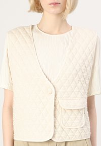Chaleco acolchado beige con escote en V, cierre de un botón y un bolsillo frontal. La tela tiene una textura suave con patrones en forma de diamante.