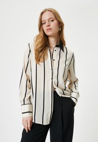Beige blouse with black vertical stripes, button-up front, classic collar, long sleeves, and black button accents. Smooth texture.