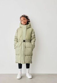 Grønne puffervest med hætte, bælte i taljen, to frontlommer og quiltet tekstur. Båret over sorte leggings og hvide sneakers.