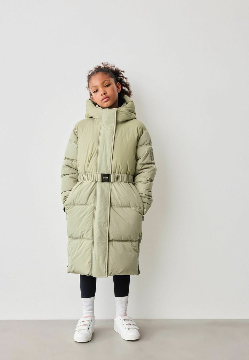 Grønne puffervest med hætte, bælte i taljen, to frontlommer og quiltet tekstur. Båret over sorte leggings og hvide sneakers.