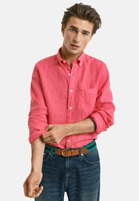 Camicia button-up rosa in lino con maniche arrotolate, taschino frontale, abbinata a jeans blu e una cintura a righe verdi. Tessuto testurizzato visibile.