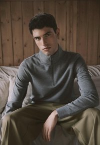 Gilet zippé gris avec un col montant, fabriqué en matériau doux. Porté avec un pantalon beige, sur fond en bois.
