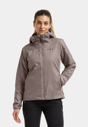 Vrouw die een lichtbruine capuchonjas met het "Didriksons"-logo en een zwarte broek draagt, staand met één hand in de zak tegen een effen achtergrond.