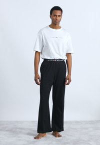 T-shirt blanc à manches courtes avec le logo "TOMMY HILFIGER" ; pantalon noir ample avec une ceinture de marque. Le modèle se tient pieds nus contre un fond clair.