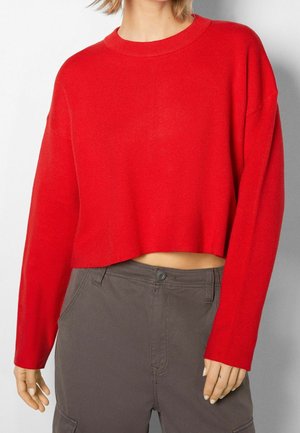 Maglione rosso a maniche lunghe con scollo rotondo, realizzato in tessuto knit; abbinato a pantaloni grigi con chiusura a bottoni.