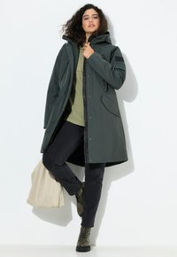Parka verde impermeabile con cappuccio, chiusura con zip e tasche laterali, indossata sopra una maglietta verde oliva chiara. Abbinata a jeans scuri e stivali oliva.