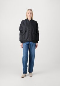 Overdimensjonert svart bomberjakke med glatt finish, ribbede mansjetter og sidelommer, kombinert med lyseblå rett-rørs jeans og beige sko.