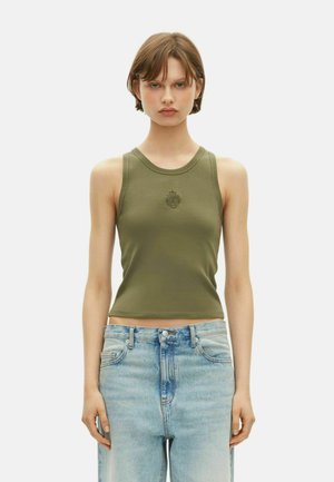 Junge Frau in einem olivgrünen ärmellosen Crop-Top mit Emblem und hoch taillierten, ausgewaschenen blauen Jeans, die vor einem schlichten weißen Hintergrund steht.
