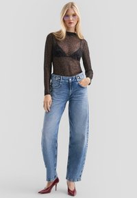 Schwarzes, durchscheinendes Longsleeve-Netzoberteil mit Leopardenmuster über einem schwarzen BH, kombiniert mit hellblauen, oversized Jeans und burgunderroten Schuhen.