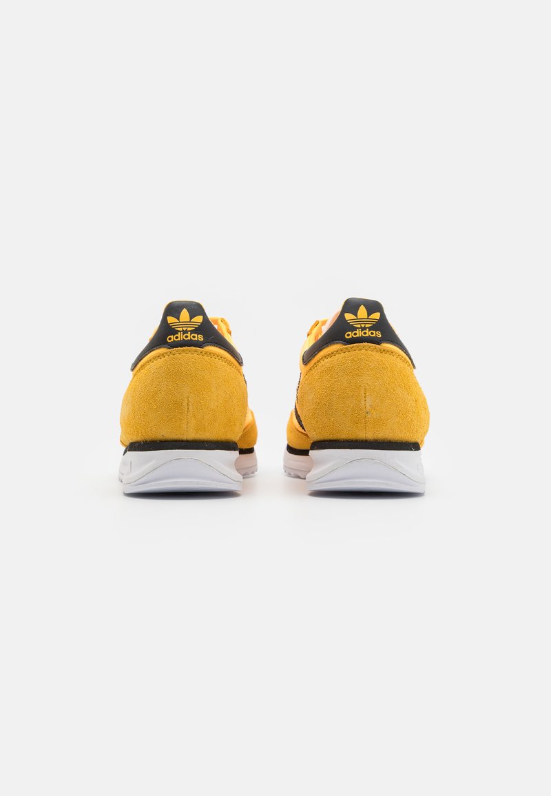 Gelbe Wildlederschuhe mit schwarzen Akzenten, ausgestattet mit einer weißen Sohle und einem schwarzen Fersensteg mit Logo. Rückansicht, die Schnürsenkel und Nähte zeigt.