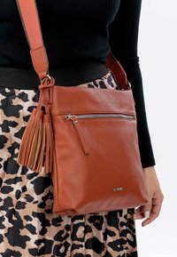 Sac en cuir brun à bandoulière avec une poche zippée et des pompons décoratifs. Surface texturée, bandoulière amovible et détail de logo sur le devant.