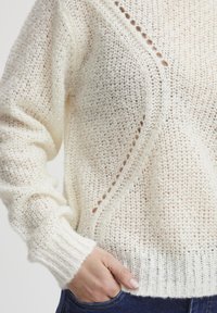 Cream knitted sweater featuring a loose, textured design, ribbed cuffs, and decorative detail on the hem with openwork patterns.