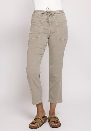 Beige high-waist pants with elastic drawstring waist and large front pockets worn with studded open-toe sandals on bare feet.