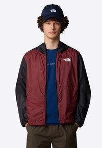 Giacca a vento rossa e nera con cappuccio, chiusura con zip e tasche con zip. Indossata sopra una maglietta blu, con dettagli del logo sul petto.