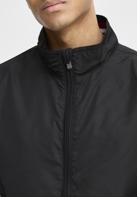 Veste légère noire avec un col montant, dotée d'une fermeture éclair frontale et d'une texture subtile. L'intérieur du col présente des rayures rouges et blanches.