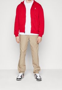 Man in een rode ritsvest, witte T-shirt, beige broek en wit, zwart en rode sneakers, staand tegen een witte achtergrond.