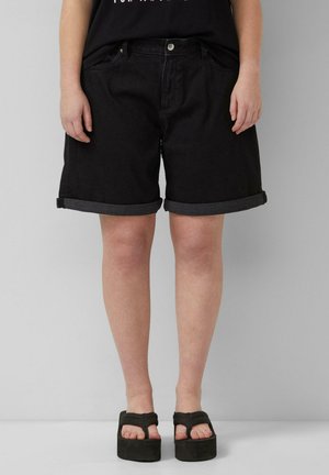 Person trägt schwarze Jeans-Shorts mit umgeschlagenem Saum, schwarze Plateausandalen und ein schwarzes Oberteil, steht vor einem einfarbigen hellgrauen Hintergrund.