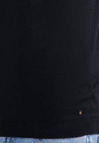 Zwarte gebreide trui met een geribde zoom. Heeft een klein, rechthoekig embleem in zwart en goud aan de linksonderkant. Eenvoudig ontwerp.