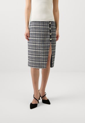 Femme portant une jupe à carreaux bleu marine et blanc jusqu'aux genoux, avec boutons à l'avant et une fente haute sur la cuisse, assortie à des talons noirs pointus.