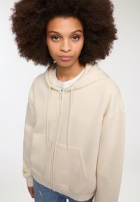 Beżowa bluza z kapturem zapinana na zamek, z podszewką w kapturze, kieszeniami z przodu i sznurkiem do regulacji. Charakteryzuje się miękką teksturą i luźnym krojem. Noszona na białej koszulce.