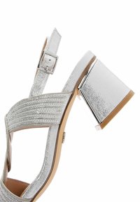 Sandale à talon argenté avec une finition pailletée, des brides texturées et un talon carré présentant des accents métalliques et un design à bout pointu.