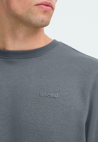 Close-up van een persoon die een grijze sweatshirt met ronde hals draagt, met het woord "blend" in reliëf op de borst.