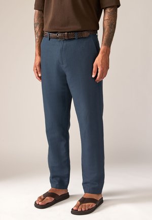 Mees, kes kannab siniseid pükse, pruuni kootud vööd, pruuni särki ja pruune sandaale, seisab nähtavate tätoveeritud kätega neutraalse tausta ees.