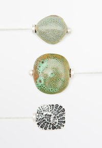 Trois perles décoratives sur des épingles en métal : une verte texturée, une brun-vert et un carré noir à motifs. Chaque perle a un accent argenté.