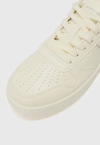 Close-up of a white sneaker featuring perforated details, lace-up closure, and a subtle logo on the side, set against a plain background.