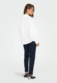Bílá oversized blůza s volnými rukávy, v kombinaci s tmavými džíny a modrými lodičkami na vysokém podpatku. Jednoduchý, moderní design.