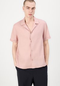 Camicia a manica corta, con bottoni, di colore rosa chiaro, realizzata in tessuto testurizzato. Presenta un colletto e una vestibilità rilassata. Abbinata a pantaloni a righe scure.