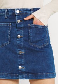 Jupe en denim bleu foncé, avec une fermeture à boutons sur le devant, deux poches latérales et des détails de couture visibles. Texture douce et design ajusté.