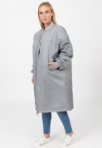 Vrouw in een lange, oversized grijze bomberjack met rits, blauwe skinny jeans en witte sneakers, staand tegen een vlakke witte achtergrond.