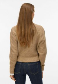 Brūns adīts džemperis ar ribotu malu, plašām piedurknēm un brīvu piegriezumu, apvienots ar tumši ziliem džinsiem, skatoties no aizmugures.