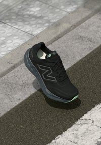 Melna New Balance skrējiena kurpe ar tumši pelēku zoli atpūšas uz asfalta blakus betona apmalei un taktilai apdarei.