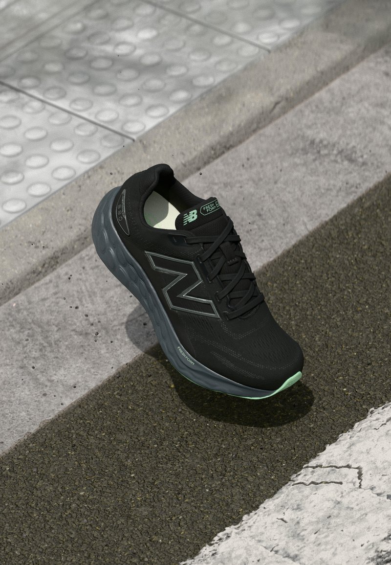 Melna New Balance skrējiena kurpe ar tumši pelēku zoli atpūšas uz asfalta blakus betona apmalei un taktilai apdarei.