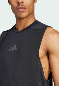 Top atlético negro sin mangas, con un diseño de cuello redondo y una textura mate. Presenta un logo reflectante en la zona del pecho.
