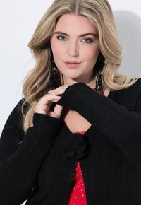 Blondýnka s vlnitými vlasy, která má na sobě černý pletený kardigan se květinovou sponou, červený top s rhinestony a dlouhé černé korálkové náušnice.