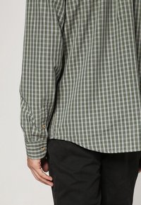 Zielono-biała koszula w kratę z długim rękawem, noszona bez wpuszczania do środka, na czarnych spodniach, zbliżenie na plecy i lewe ramię.