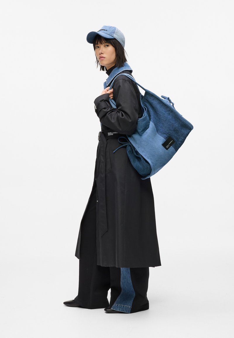 Γυναίκα με μαύρο παλτό και παντελόνι patchwork κρατά μεγάλη μπλε τσάντα σε δύο τόνους και φοράει τζιν καπέλο που ταιριάζει, μπροστά σε άσπρο φόντο.