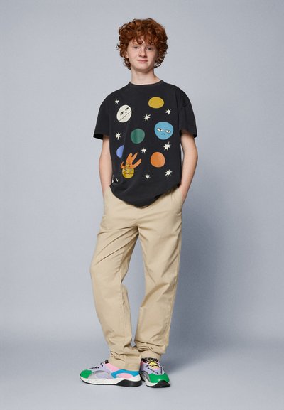 Črna grafična majica s pisanimi nebesnimi motivi, bež hlače in večbarvni športni copati iz mrežaste tkanine z drznimi poudarki.