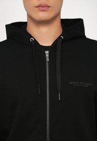 Svart zip-up hettegenser med snor, med sølvfarget glidelåsdetaljer og en teksturert logo "Armani Exchange" på brystet.