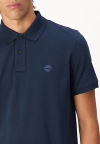 Tamsiai mėlynas polo marškinėlis su smailiu apykakliu, trimis mygtukais, mažu logotipu ant krūtinės ir tekstūruota medžiaga. Trumpomis rankovėmis.