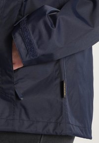 Manche de veste bleu marine avec bride Velcro, main dans la poche latérale, et petite étiquette noire cousue près de l'ourlet.
