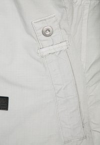 Jasnoszara tkanina o subtelnej siatkowej strukturze, z zapięciem na zatrzask oraz czarną naszywką na lewym boku.