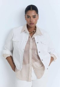 White denim jacket with two front pockets, button closure, and rolled sleeves, worn over a beige striped shirt. Simple design, neutral colors.